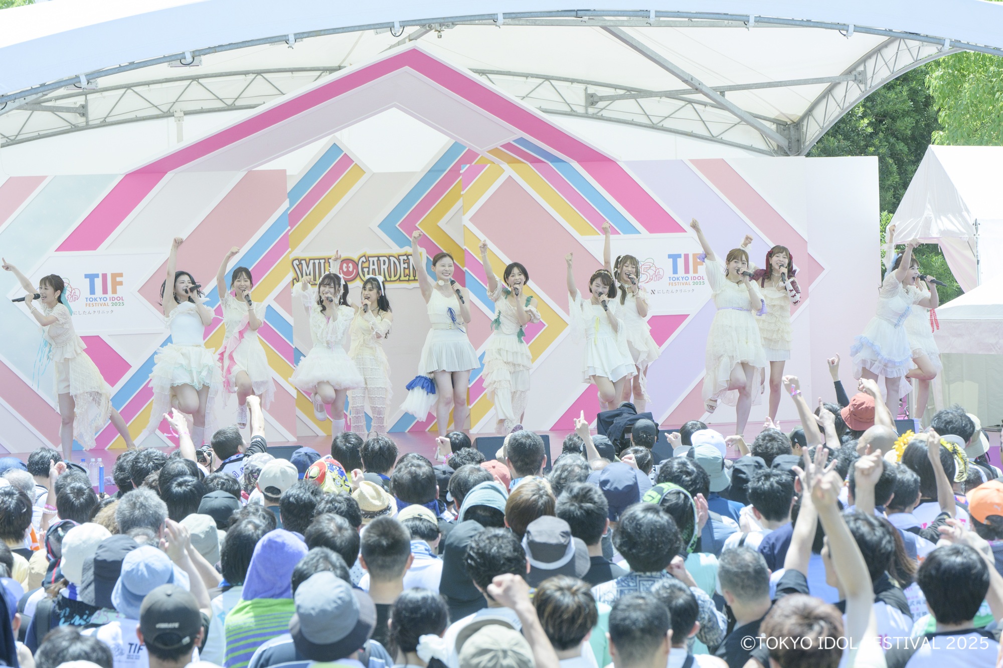 アイドルグループがパフォーマンスを行うステージの様子。観客は大勢集まり、楽しそうに手を挙げている。背景にはカラフルな装飾が施された舞台が見える。