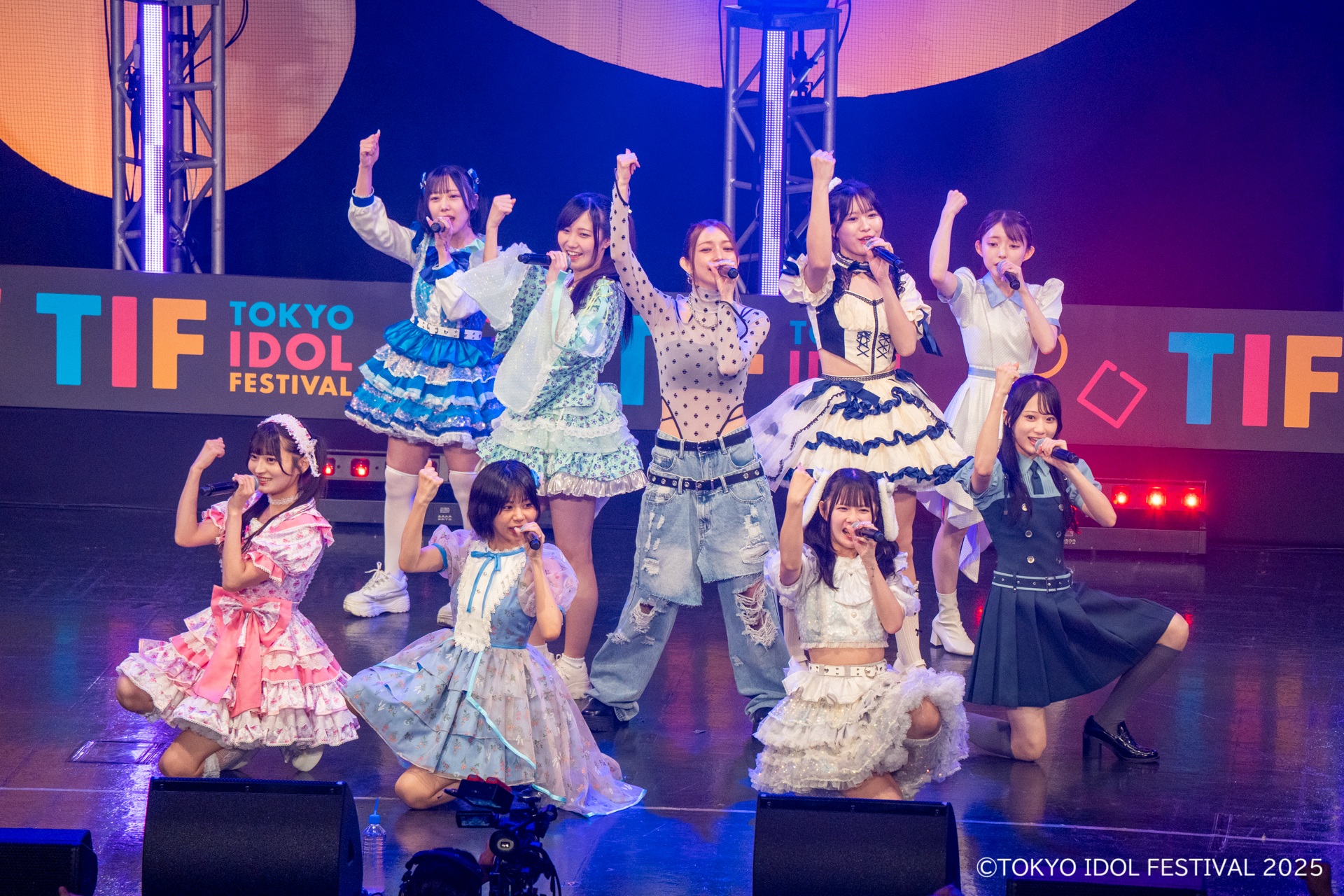 ライブパフォーマンス中のアイドルグループ、カラフルな衣装を着たメンバーがステージで盛り上がっている。背景には「TOKYO IDOL FESTIVAL」のロゴが見え、観客の前で熱意を示している様子。