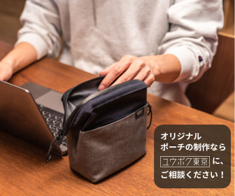 デスク上でノートパソコンを使っている人の手元に置かれたオリジナルポーチ。ポーチはグレーと黒の組み合わせで、開いている状態。左上には「オリジナルポーチの制作ならユウボク東京に、ご相談ください！」というテキストが表示されている。