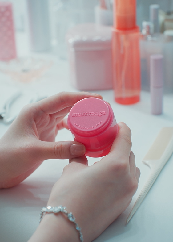 手に持たれたピンク色のヘアスタイリングスティック「matomage」が映っている。背景には他のヘアスタイリング商品が並ぶ。