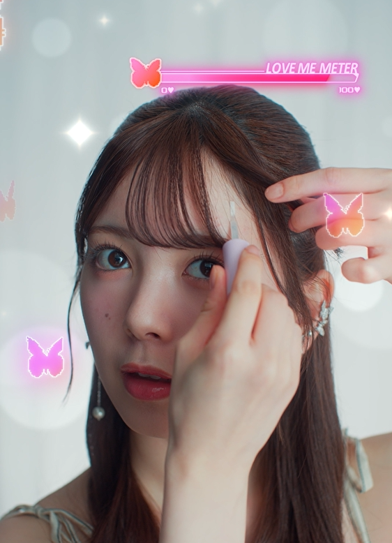 A young woman is styling her hair with a tool, focusing on her bangs. The background features decorative butterflies and a 'LOVE ME METER' graphic indicating her confidence level. The atmosphere conveys a blend of concentration and positivity.