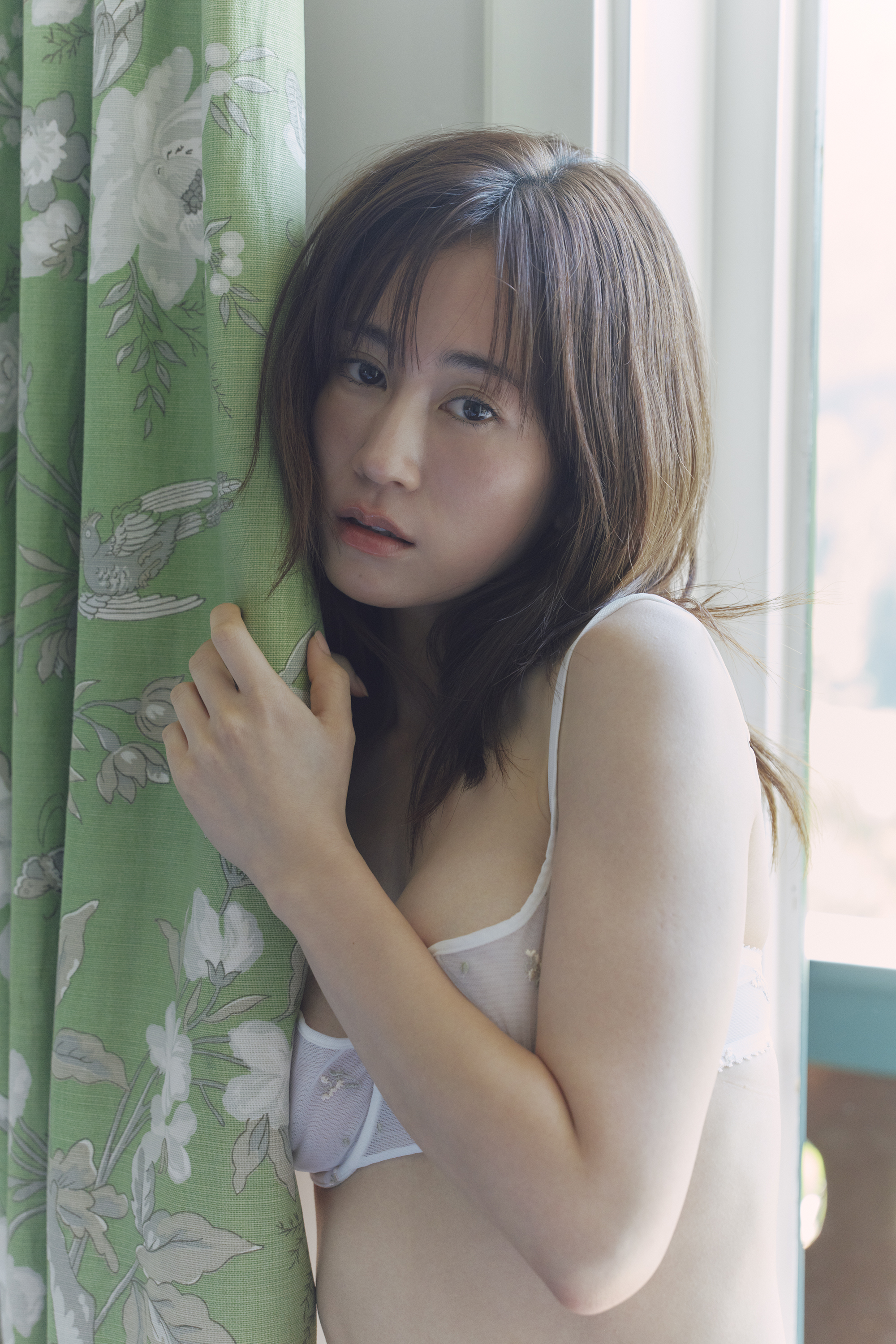 女性が緑のカーテンのそばに立っている姿。彼女は白いランジェリーを着用し、柔らかい表情を浮かべている。