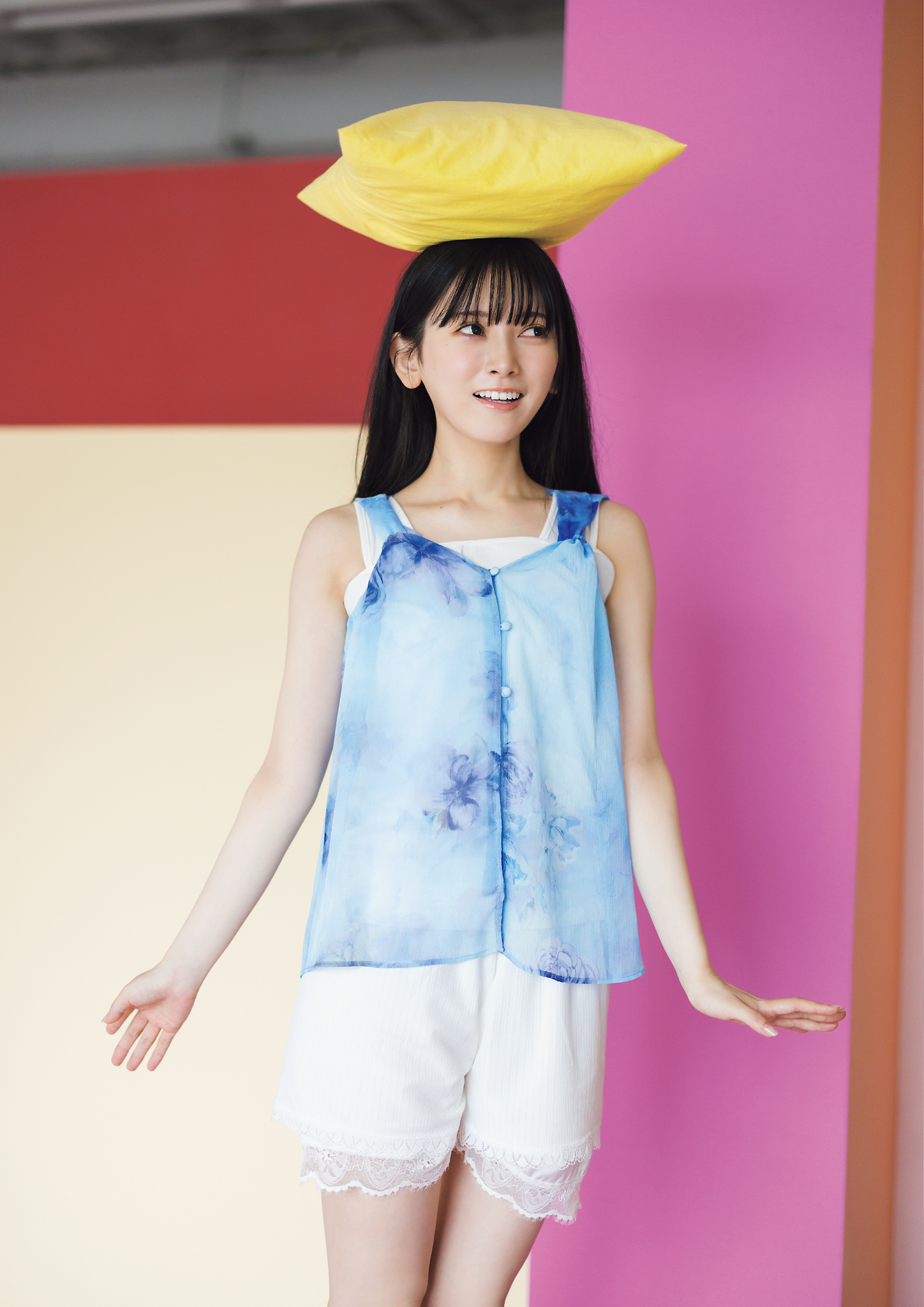 青い花柄のトップスと白いショートパンツを着た女性が、黄色のクッションを頭に載せて微笑んでいる。背景にはピンクとベージュのカラフルな壁がある。