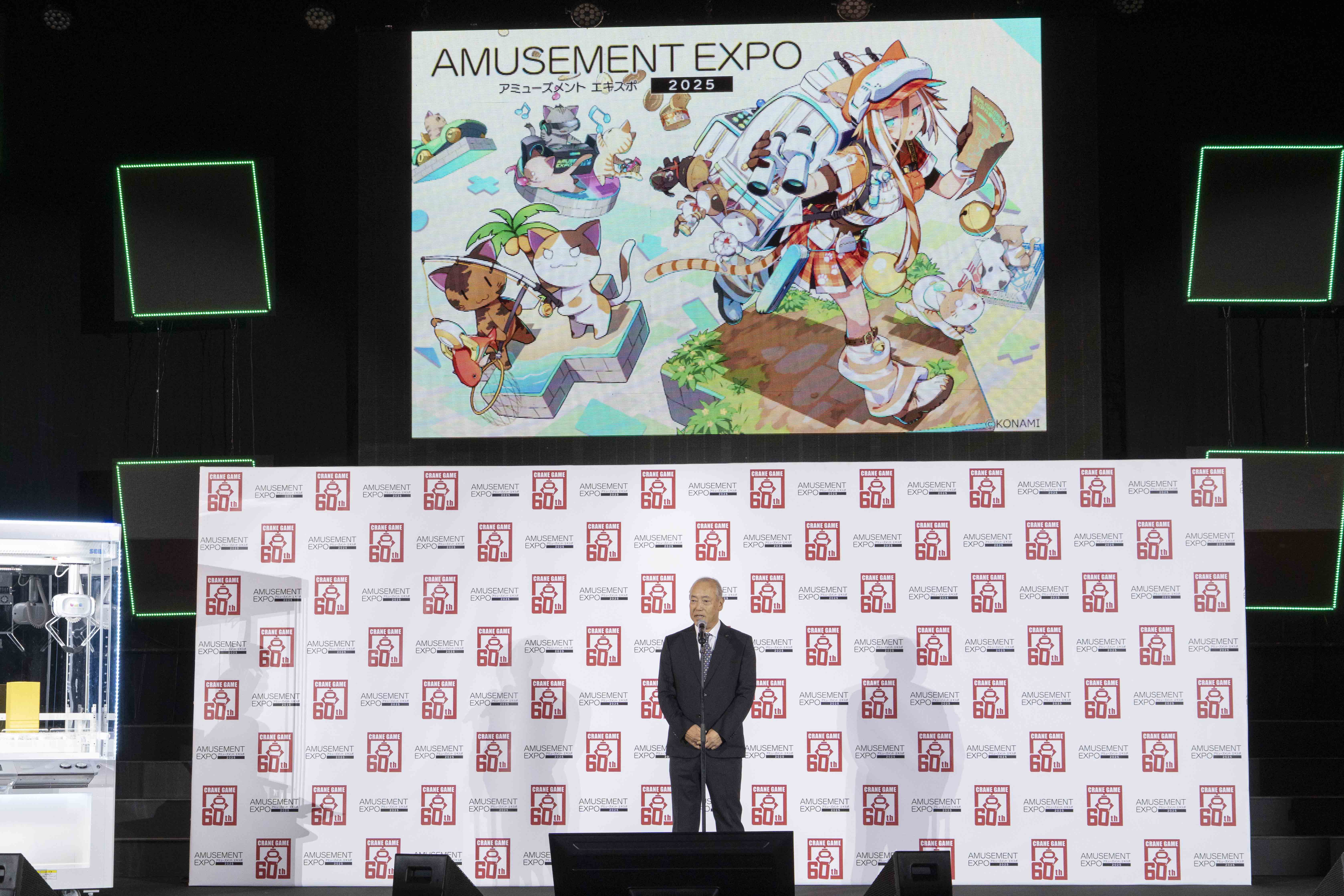 アミューズメントエキスポ2025のオープニングセレモニーで登壇する男性が、ステージ上でスピーチを行っている様子。背景にはカラフルなキャラクターアートが描かれたスクリーンが表示されている。