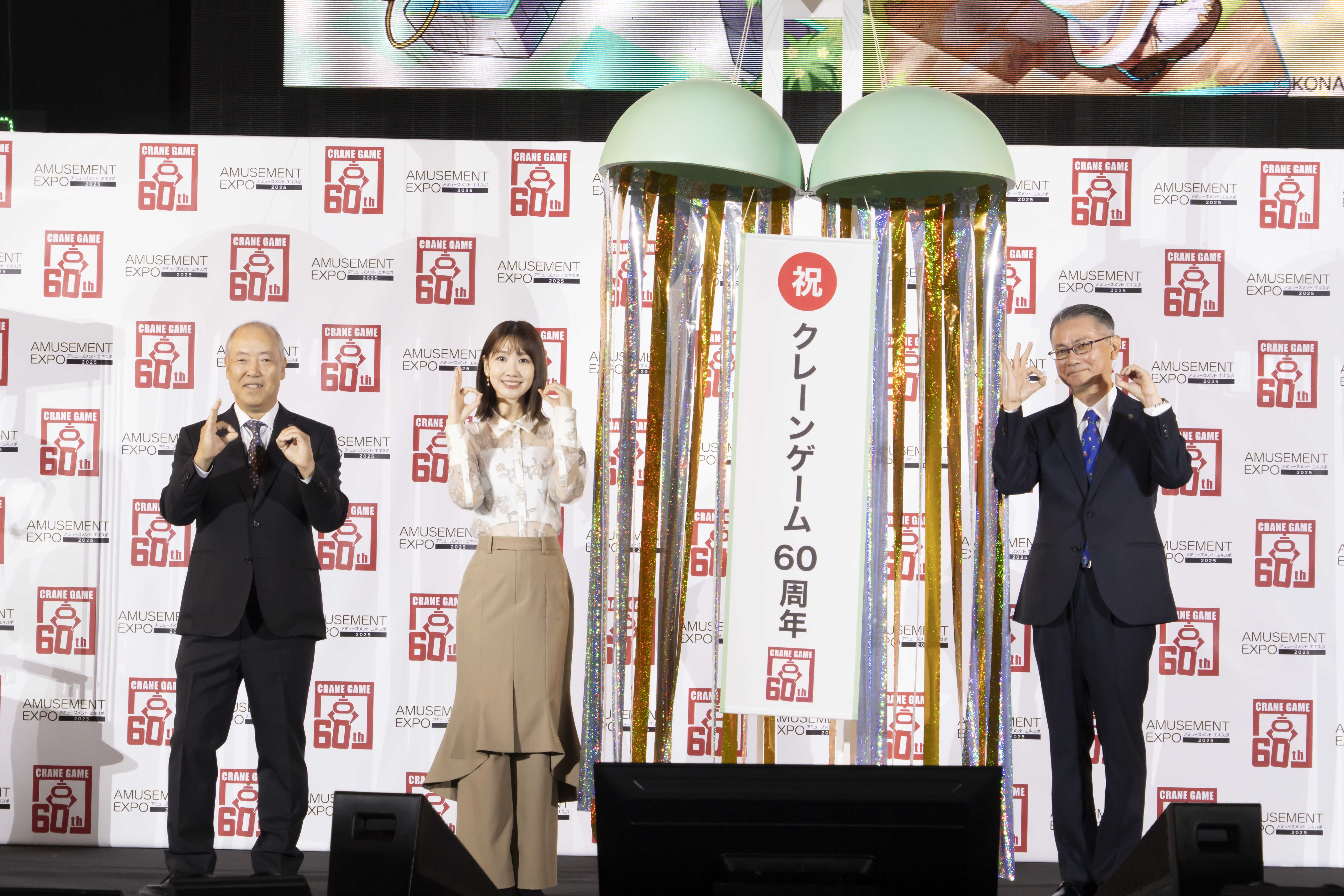 クレーンゲーム60周年記念セレモニーで、柏木由紀と他の出演者が祝福のポーズを取っている様子。背景にはアミューズメントエキスポのロゴが見える。