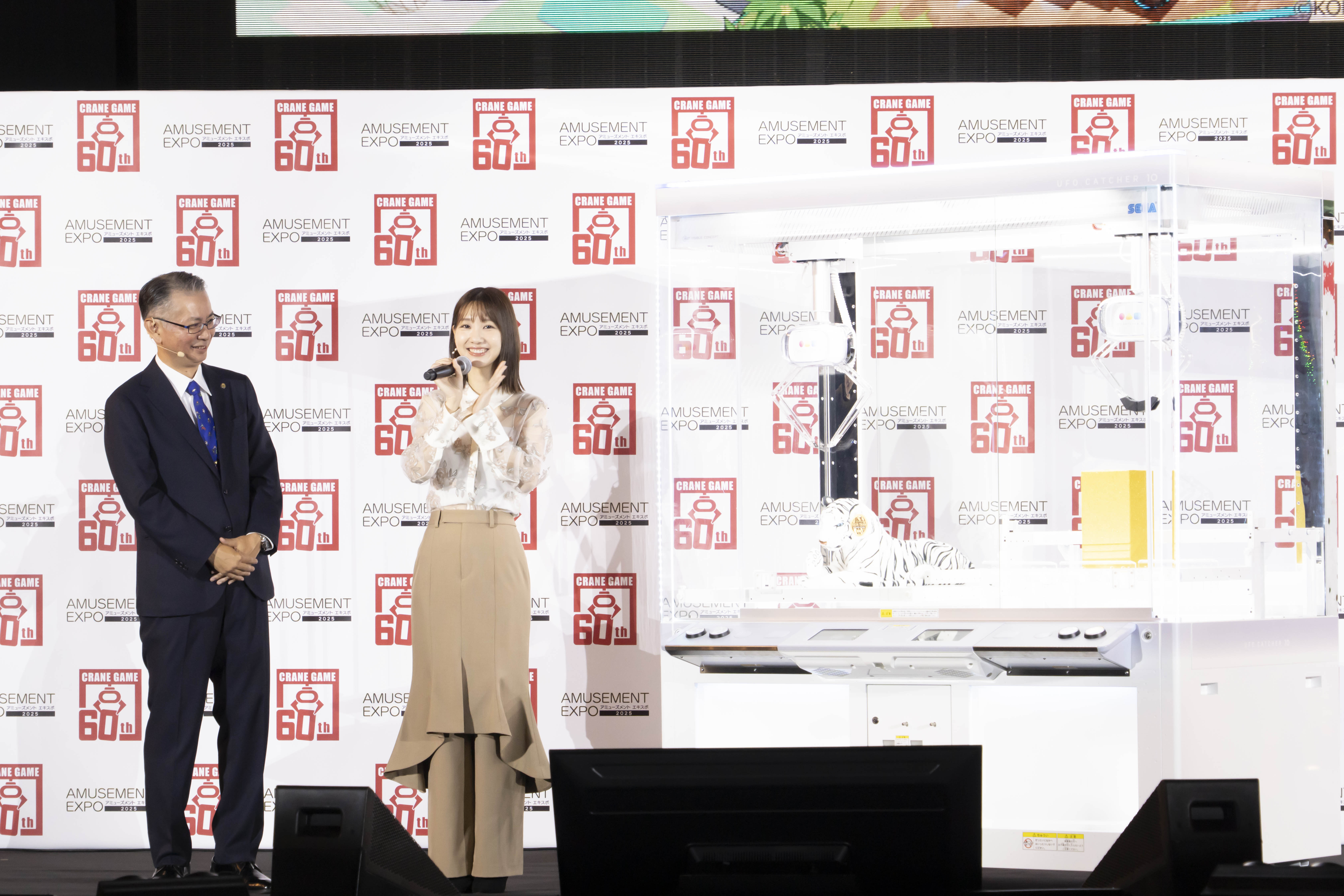 タレントの柏木由紀が「アミューズメントエキスポ2025」のオープニングセレモニーでマイクを持って笑顔で話している様子。背景にはクレーンゲームの60周年を祝うロゴが見える。