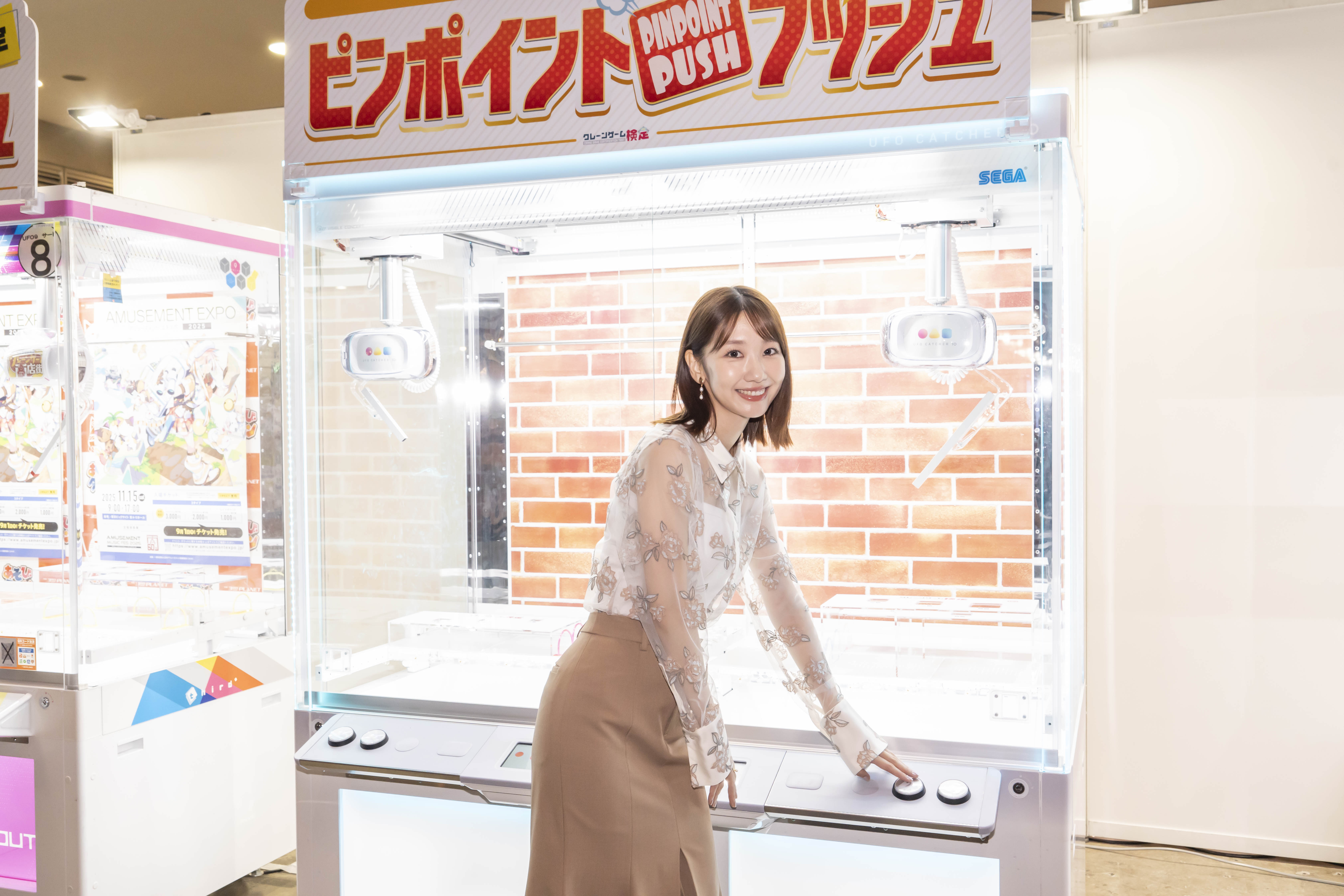 タレントが最新のクレーンゲーム『ピンポイントプッシュ』で遊んでいる場面。ゲーム機の前で微笑みながらボタンを操作する姿が写されている。
